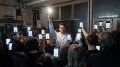 In the photo: a man is holding a lit light bulb, and a group of people are holding mobile phones