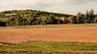 In the photo: a rural landscape: in the foreground, a field; in the background, a tractor, a forest, and a house.