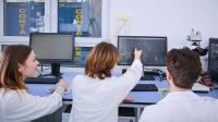 In the photo, three people are sitting at a computer, and one is pointing at the screen